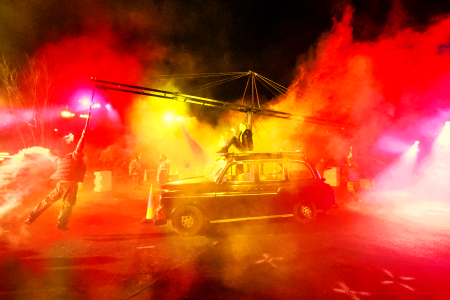 An aerialist sits on top of a taxi cab, with another performer suspended from a long aerial bar attached to the taxi. There is orange and yellow stage light and smoke covering the whole mage from Rave New World.
