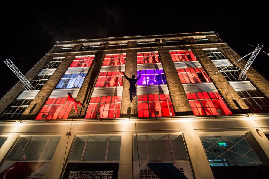 A shot of the hat factory arts centre in Luton with lit up red and purple windows and aerialists scaling the walls