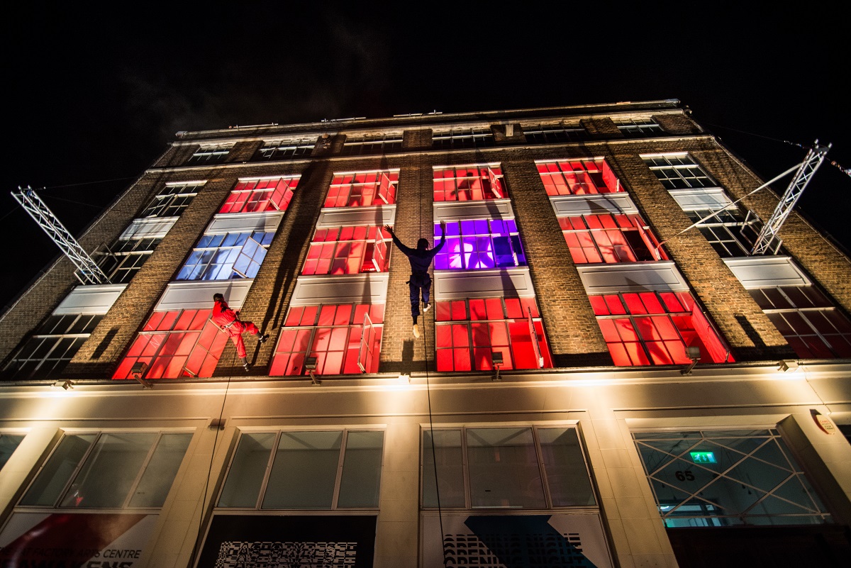A shot of the hat factory arts centre in Luton with lit up red and purple windows and aerialists scaling the walls