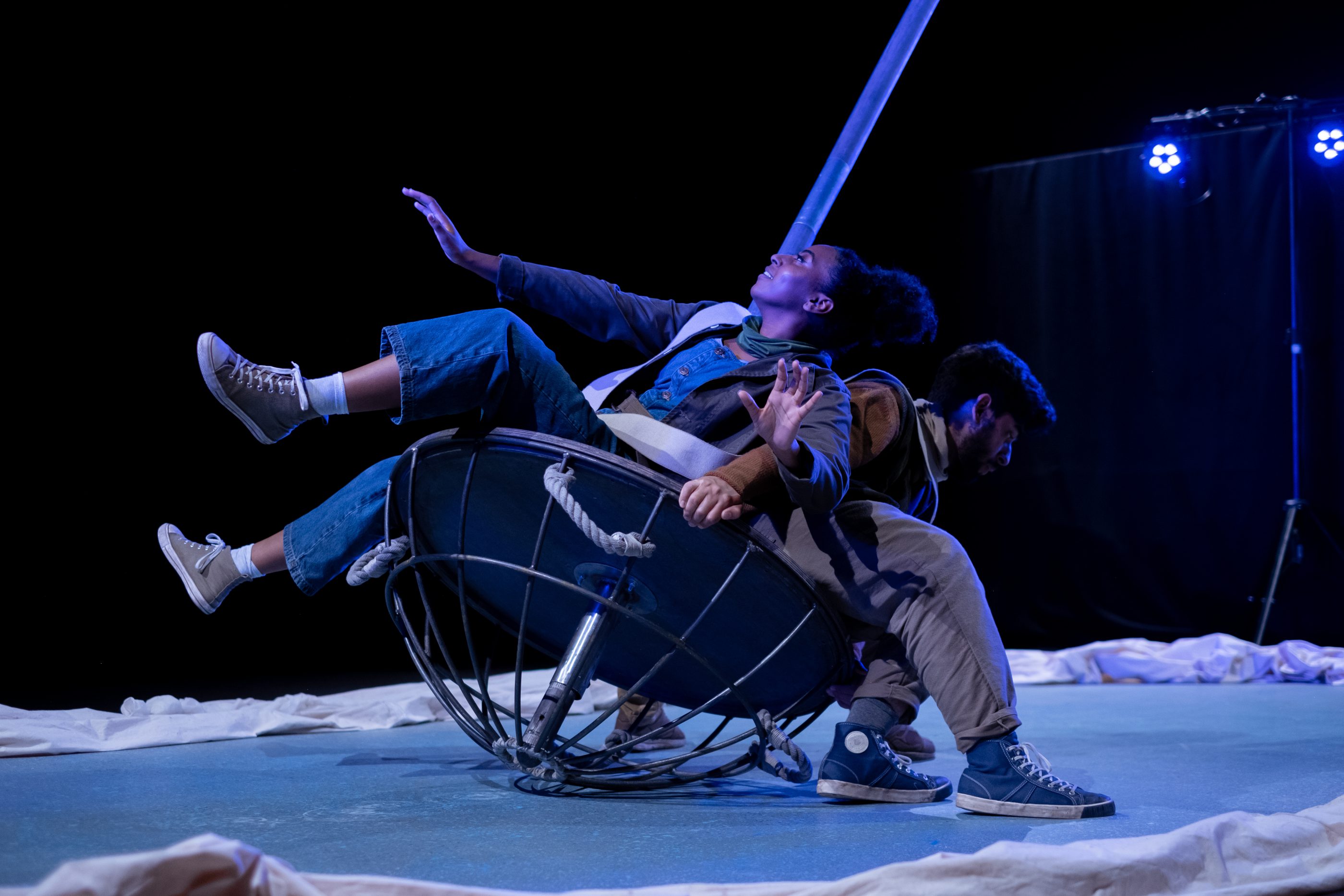 Three performers lying on a piece of set, with a large pole, which gives the impression of a boat amongst a simple set with a white floor and black background