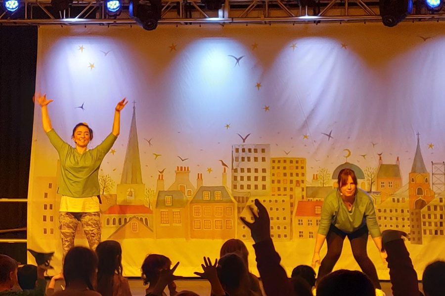 Two Mindfulness project team in yoga poses onstage with a city landscape backdrop in the background. There are an audience of children with hands up in the air, following the poses.