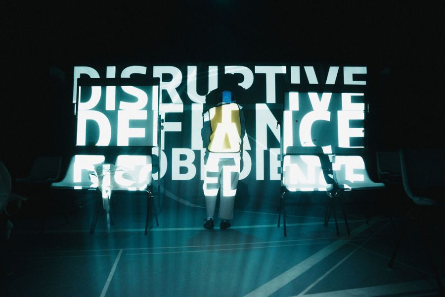 A performer standing onstage with large letters projected over them and the back screen. The words say Disruptive Defiance Disobediance