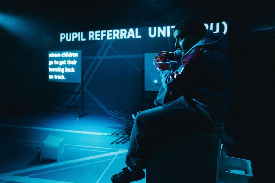 A performer sitting in the forefront with a backdrop projection saying Pupil Referral Unit.
