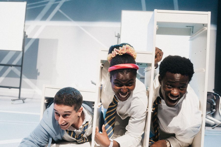 Three performers pushing themselves inside a white shelving unit that is shaped like stairs. They look happy and like they are having fun