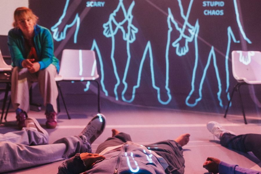 A performer lying down on the floor and another performer in the background sitting on a chair leaning forward. Behind them are projections of body outlines with a number of words projected inside them.