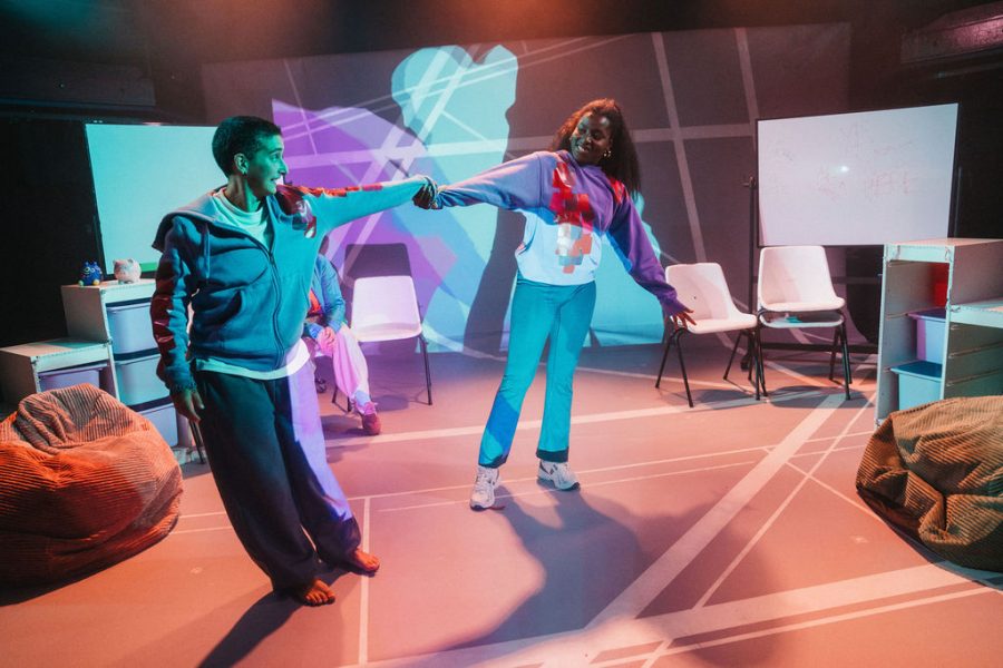 Two performers joined by the hands smiling at each other, with a colourful back wash of colour and projections of a blurry body image on the backdrop.