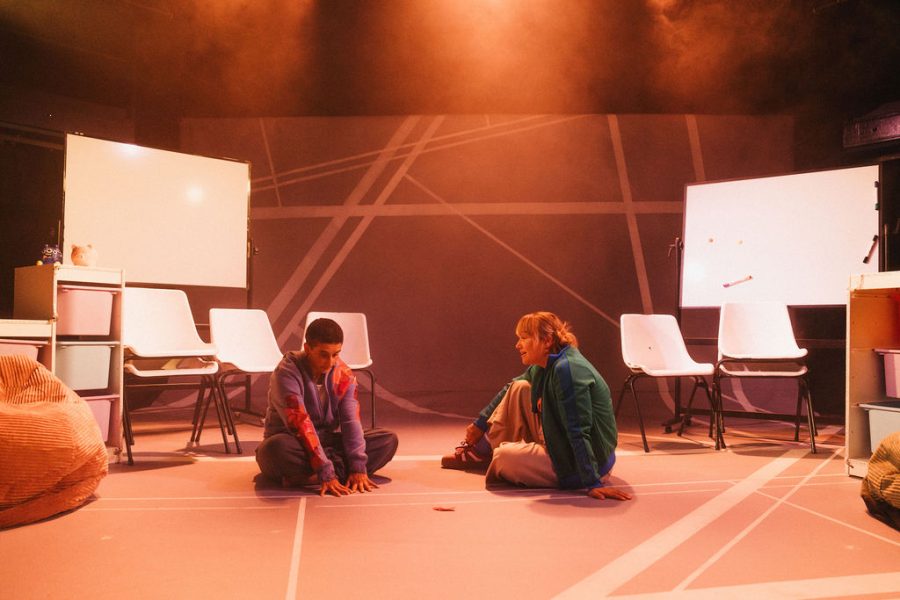Two performers sitting with legs crossed facing towards each other. There are 2 whiteboards and some white school chairs and white shelving units plus a bean bag.