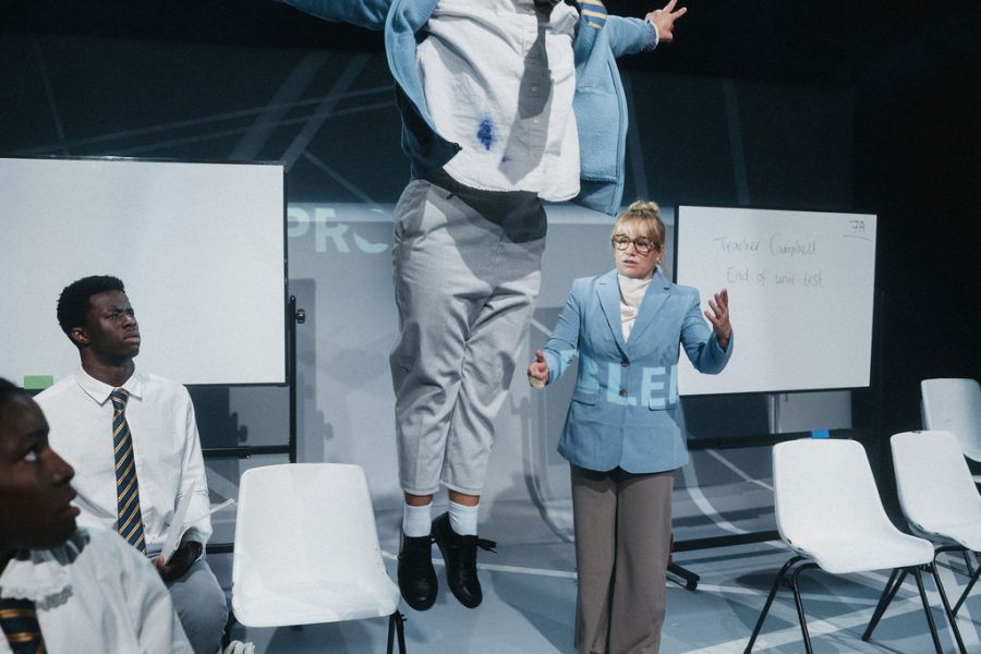 A performer jumping high up in the air and the others looking on amongst the set with white school chairs and 2 whiteboards in the background