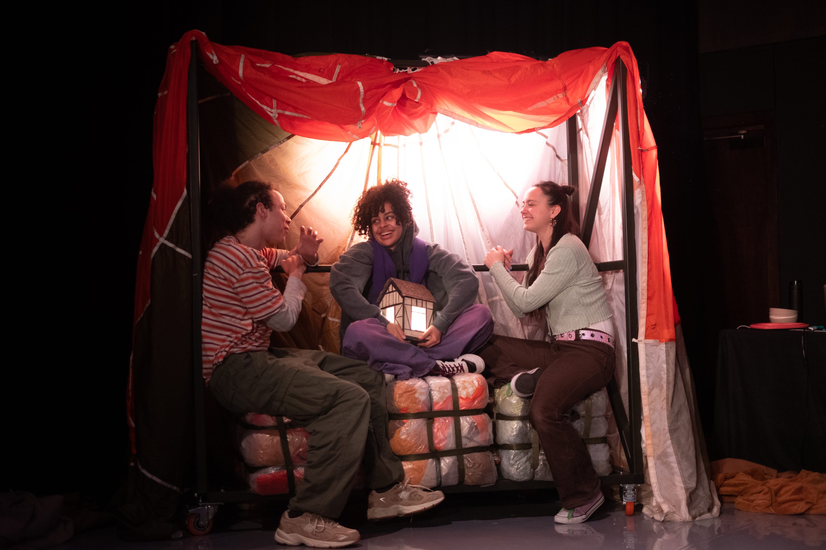 Three perfomers sit on a set in conversation