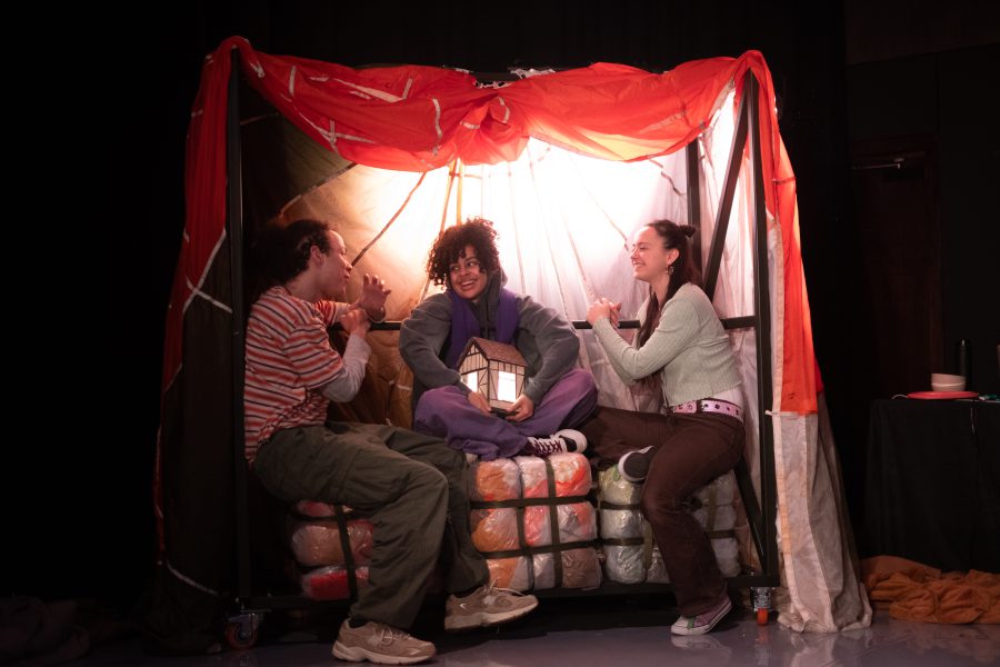 Three perfomers sit on a set in conversation