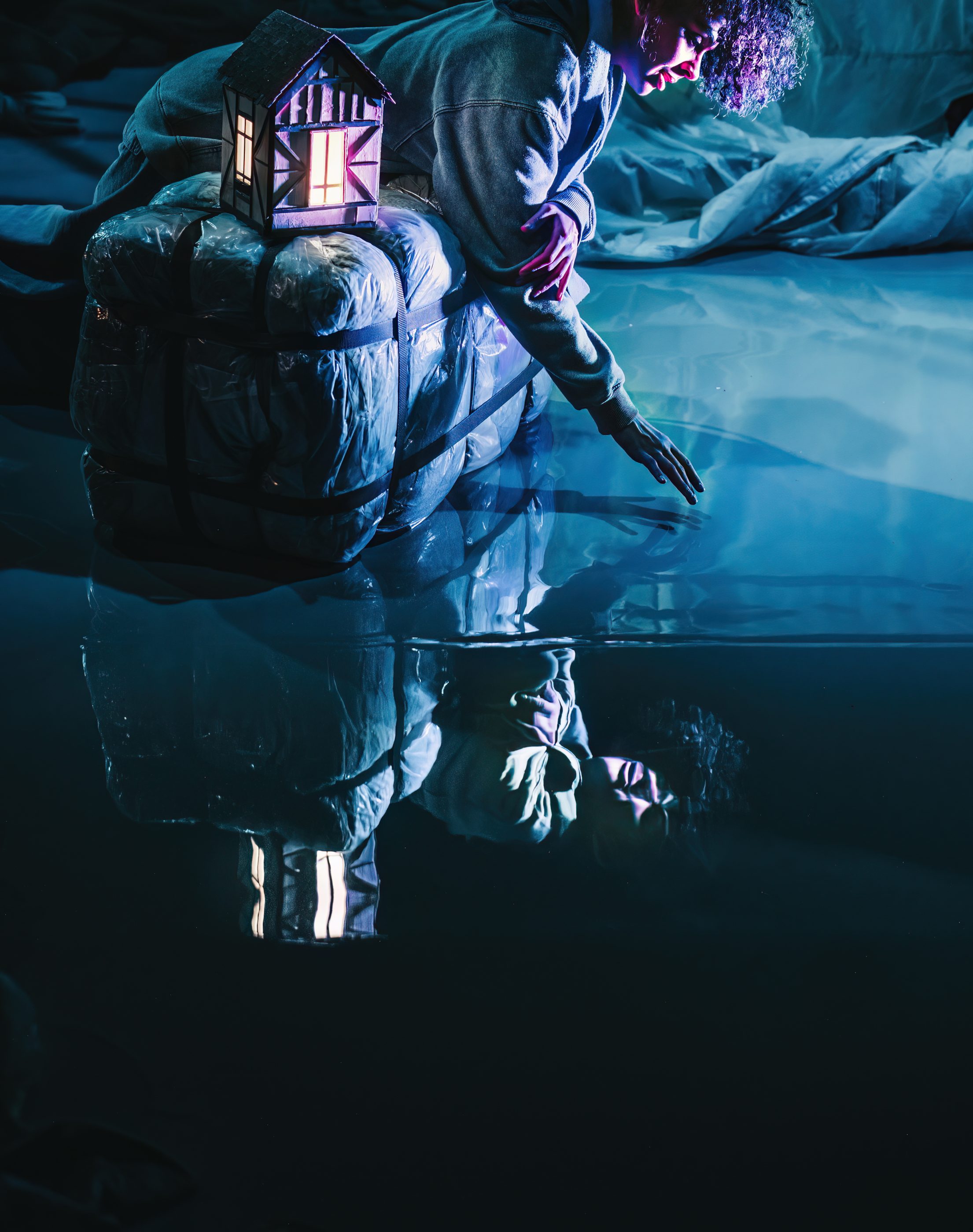 A person sitting next to a tiny glow up house model, reaching into a floor, which looks like water and shows a reflection of the person and the little house.