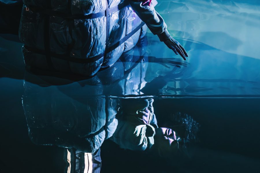 A person sitting next to a tiny glow up house model, reaching into a floor, which looks like water and shows a reflection of the person and the little house.