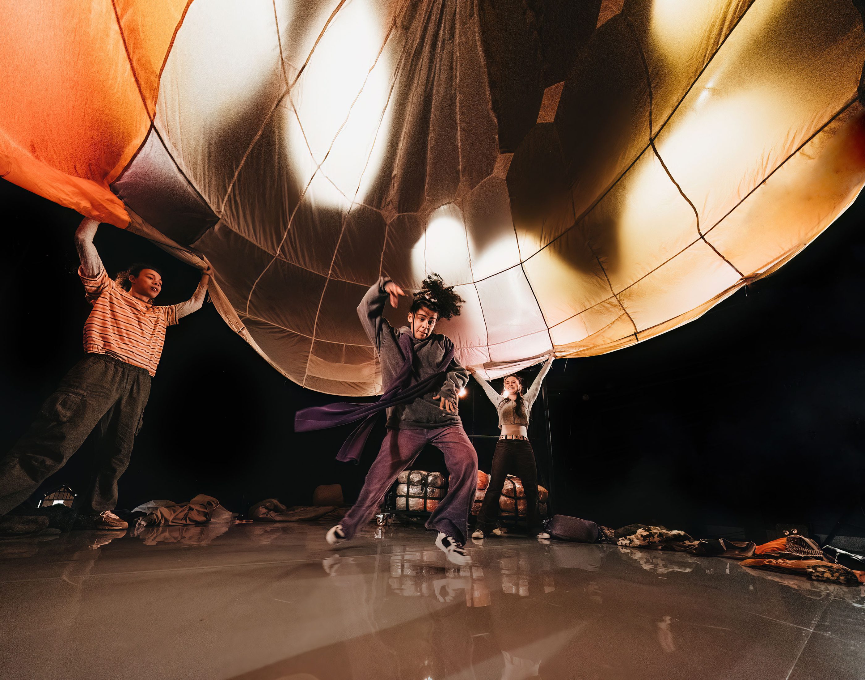 Three performers in the production Belongings under an orange, brown and cream parachute, one performer dances underneath it whilst the other two hold it up, upon a reflective looking floor.