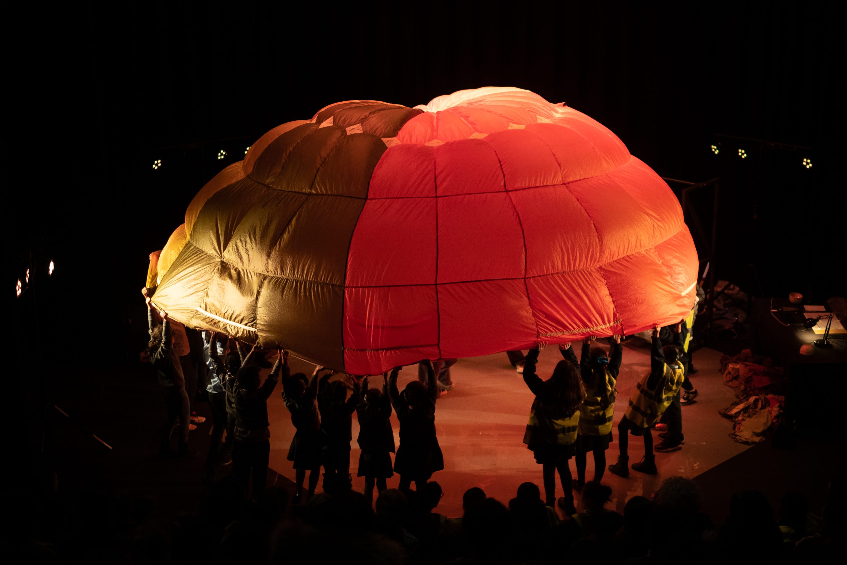 A group of young people holding up a large orange parachute onstage