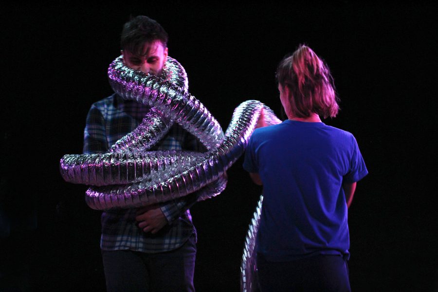 A performer with a long silver tube attached to his arms which is wrapped around his body. Another performer stands, we see the back of their head and they are holding the silver tube.