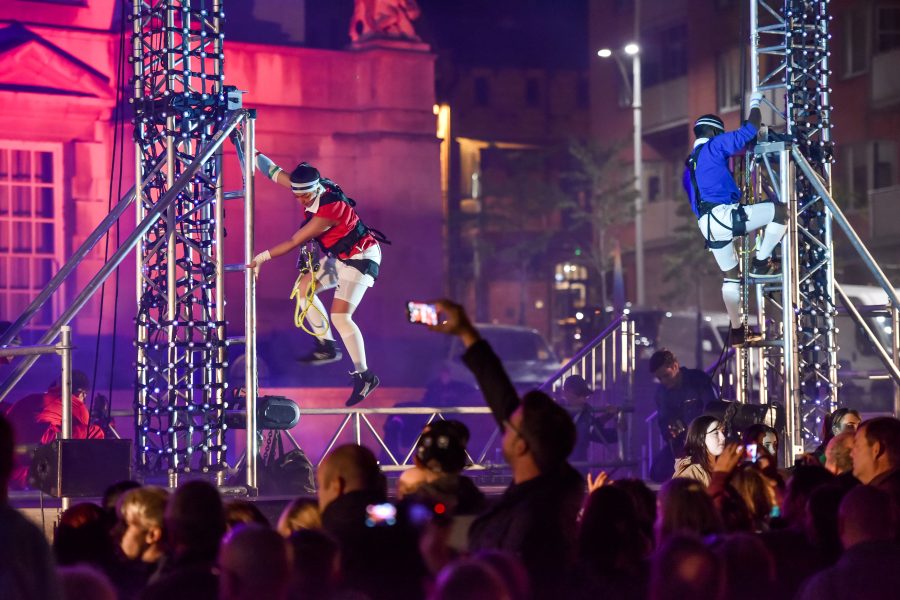 Two performers climb a scaffolding in a night time production, with a large audience watching and filming the action.
