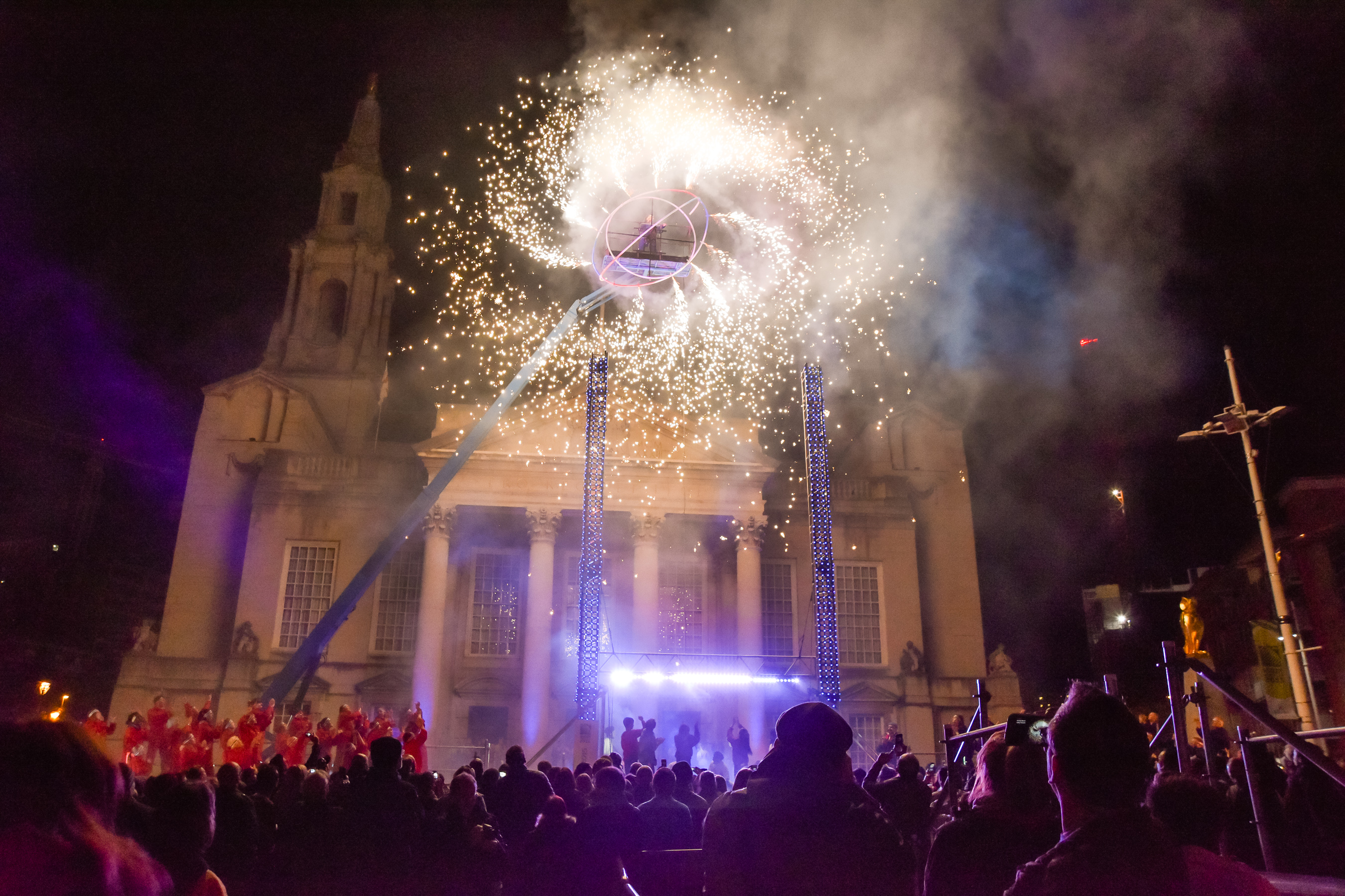 A Catherine wheel with performer inside suspended high in the air outside a large histiric building with large audience watching and lots of ensemble performers standing underneath