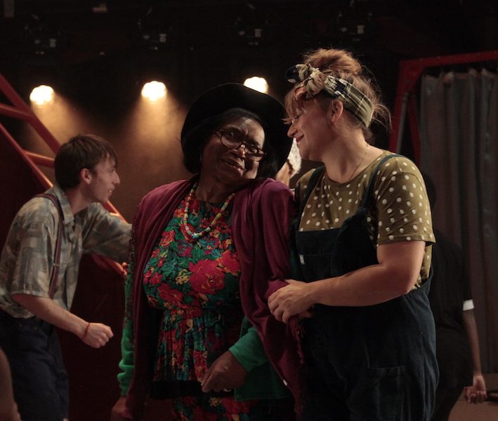 Two performers link arms and look at each other smiling on the set of Half Life, with a performer in the background moving behind them