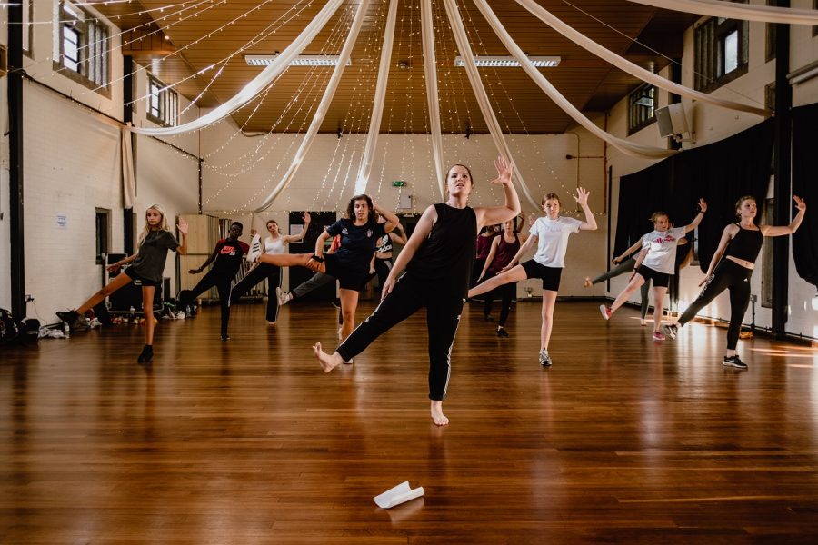 A group of dancers from NGYT, in a dance pose, one hand outstretched, balancing on one leg, in a large rehearsal space with a wooden floor and ribbon cloth drapes hanging from the ceiling