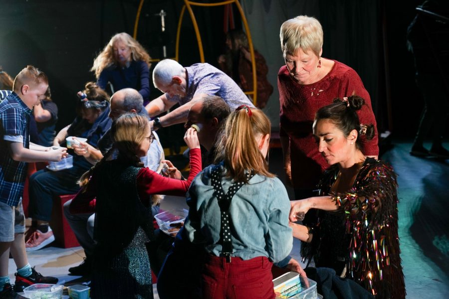 A group of an ensemble made up of all ages, sitting in a row applying face paint and helping each other.