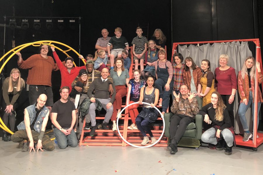 The intergenerational ensemble of Half Life in Stockton, sitting on the set which includes a large yellow german wheel, red scaffolding climbing frame and a red screen on wheels. There are over 30 people pictured of all ages.
