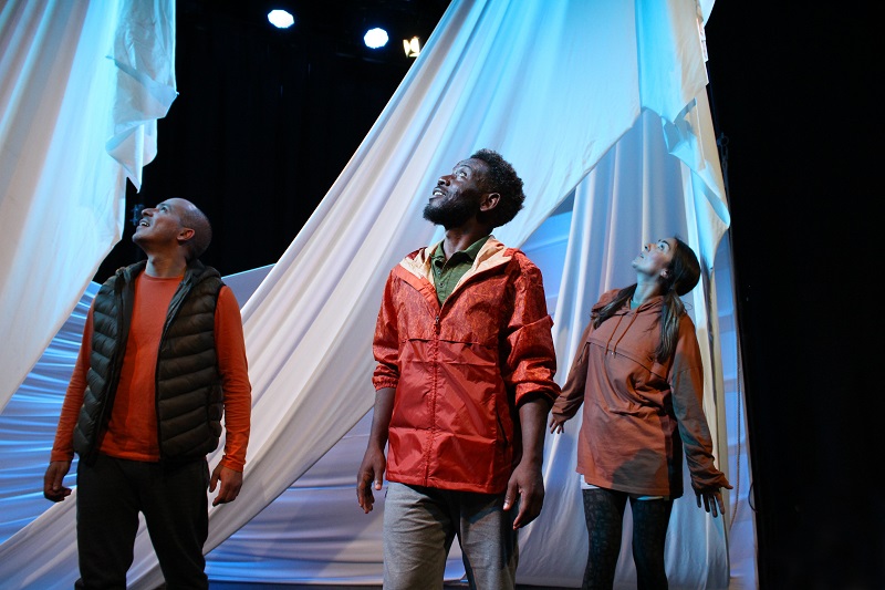 Three performers onstage looking up. They are surrounded by white drapes and wear orange costumes.