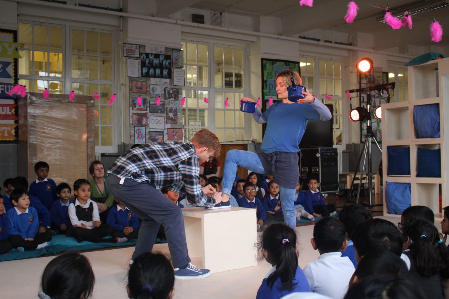 A scene from Need A Little Help with two performers surrounded by schools audience watching the action.