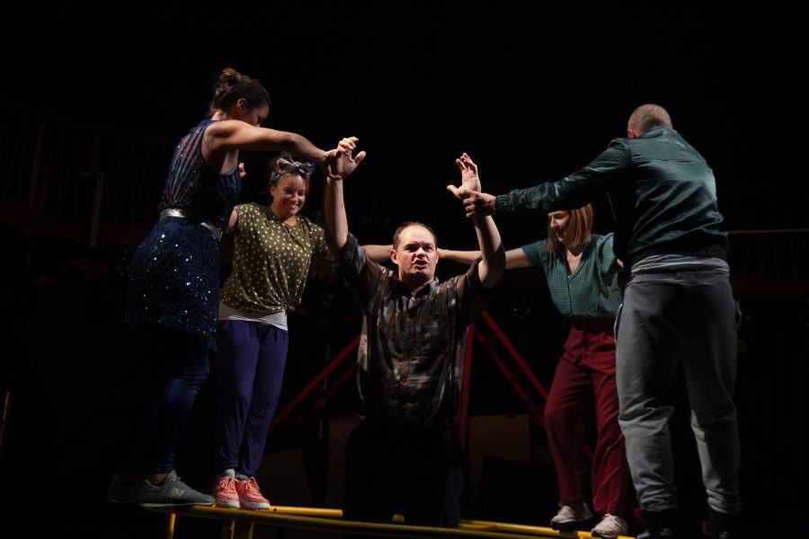 A group of performers standing on a yellow German Wheel which lies flat on the floor. Another performer stands in the middle with arms in the air holding hands with two of the performers. The image is from Half Life.
