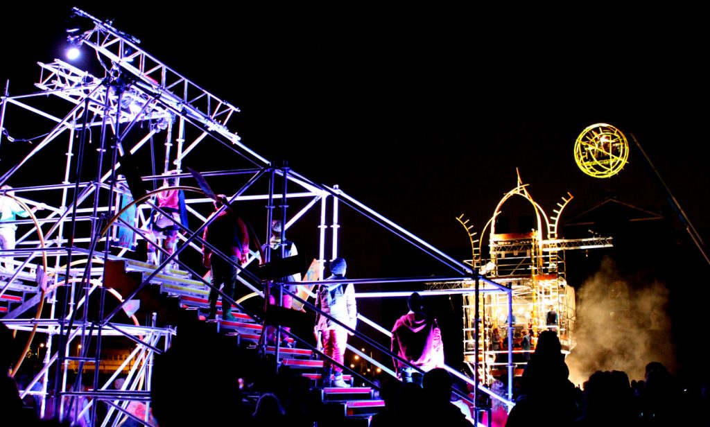 A scaffolding structure with performers standing on steps on it. It is night time and the scafflding is well lit up looking at another large scaffolding tower with a Yellow Catherine Wheel suspended from it. A large outdoor show attended by hundreds of people in the audience (some audience back of heads are pictured)