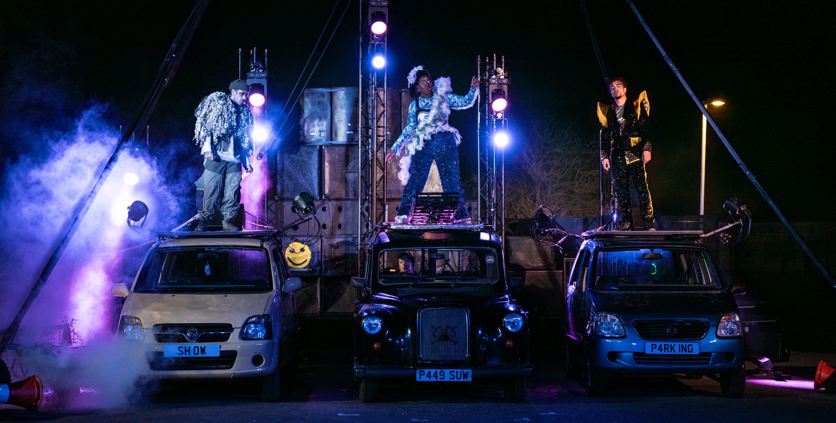 Three performers stand on top of 3 cars with stage lights and a sound system, behind them.