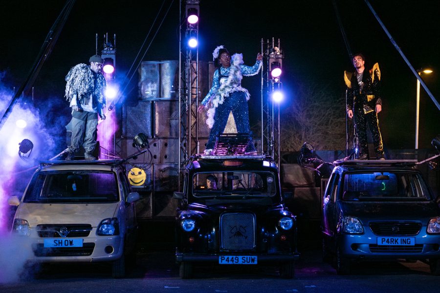 Three performers stand on top of 3 cars with stage lights and a sound system, behind them.