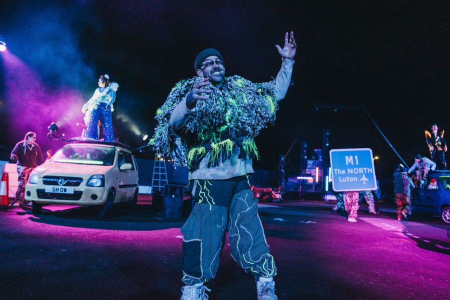 A performer in an oversized waistcoat made with multiple layers of yellow, green and grey cloth and baggy trousers and a beany hat stands at the forefront of the image with arms in the air. Behind them to the right is a sign being held up that says M1 North Luton. To the left back of image another performer stands on top of a yellow car. Taken from Rave New World in Luton.
