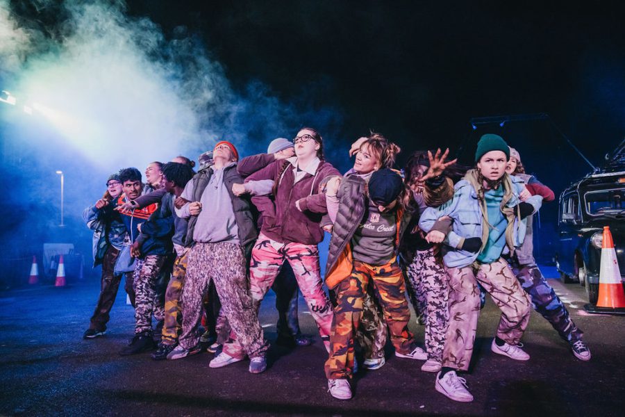 A large group of performers clumped together linking arms, wearing mutli-coloured costumes and beany hats, with a smoky stage lit background, from Rave New World.