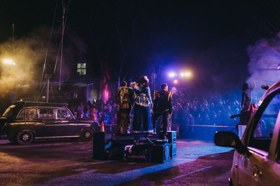 Three performers looking out to an audience, standing on barrels with a car in the forefront and a taxi cab in the left of the image. There is a purple/blue stage light washing over the scene, taken from Rave New World.