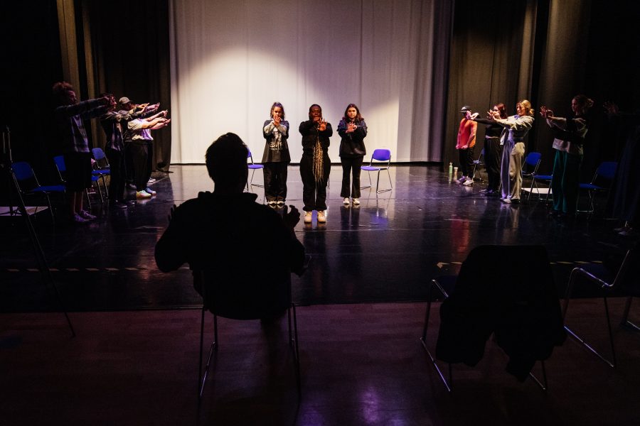 Working on a scene in a drama studio in a college. The director watches from audience as performers onstage are in a number of physical poses with their arms outstretched.