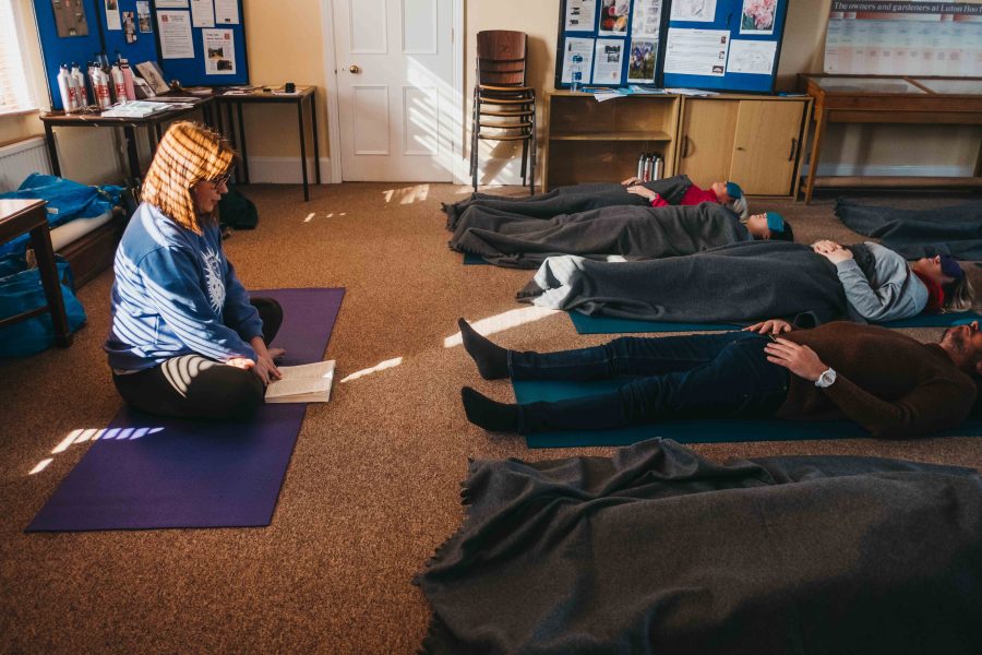A yoga practitioner reads out a meditation to a group of adults in a Mindfulness for Business session. There is sunlight rays coming through some blinds and a group of adults are lying down on yoag mats, some with blankets over them and some with eye pillows too.