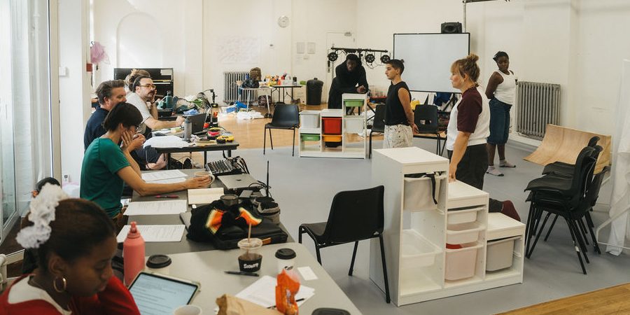 A rehearsal room with people standing on a stage area and other people sitting at tables looking towards them, There are a lot of props and set dotted around in a well lit white washed room.