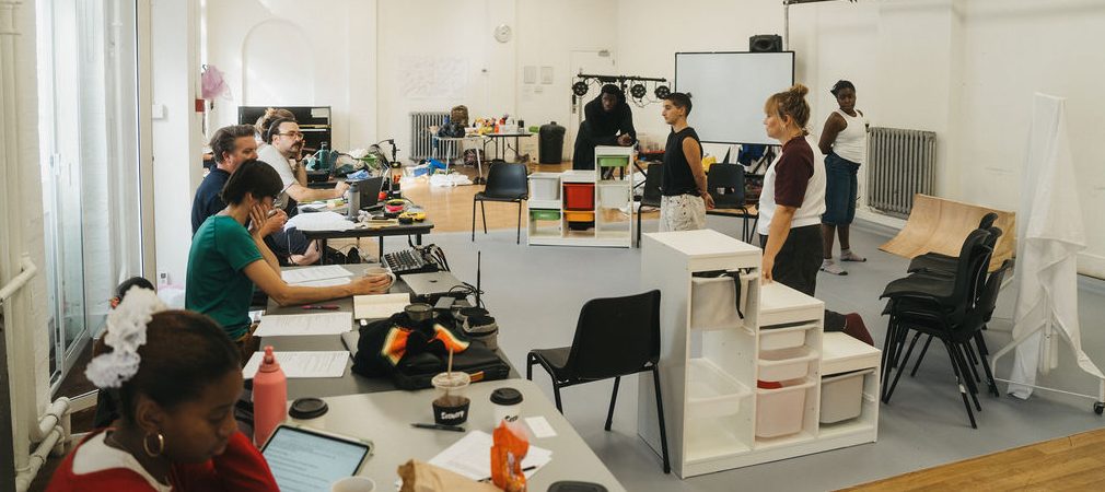 A rehearsal room with people standing on a stage area and other people sitting at tables looking towards them, There are a lot of props and set dotted around in a well lit white washed room.
