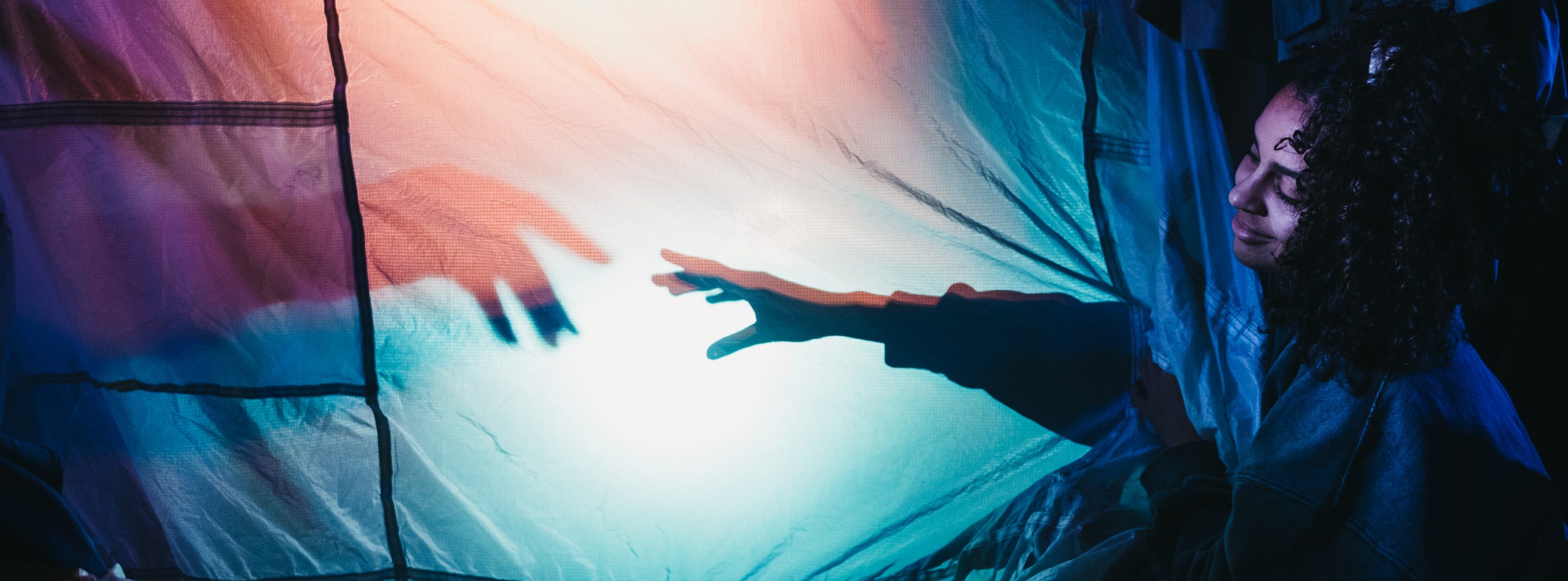 A perfomer smiles looking at their arm which reaches behind a lit up white sheet, creating a shadow, reaching towards another hand coming in from the left hand side.