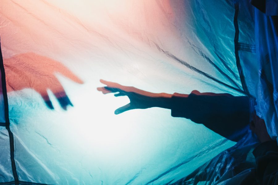 A perfomer smiles looking at their arm which reaches behind a lit up white sheet, creating a shadow, reaching towards another hand coming in from the left hand side.