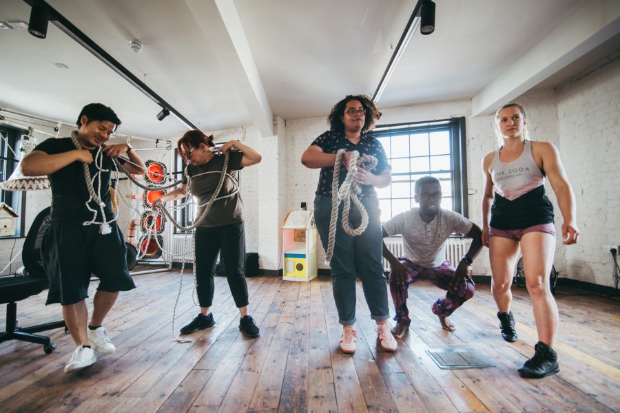 5 Performers standing and crouching, some are holding rope, in a large open well lit rehearsal space. There are speakers in the background and a large window with natural light coming in.