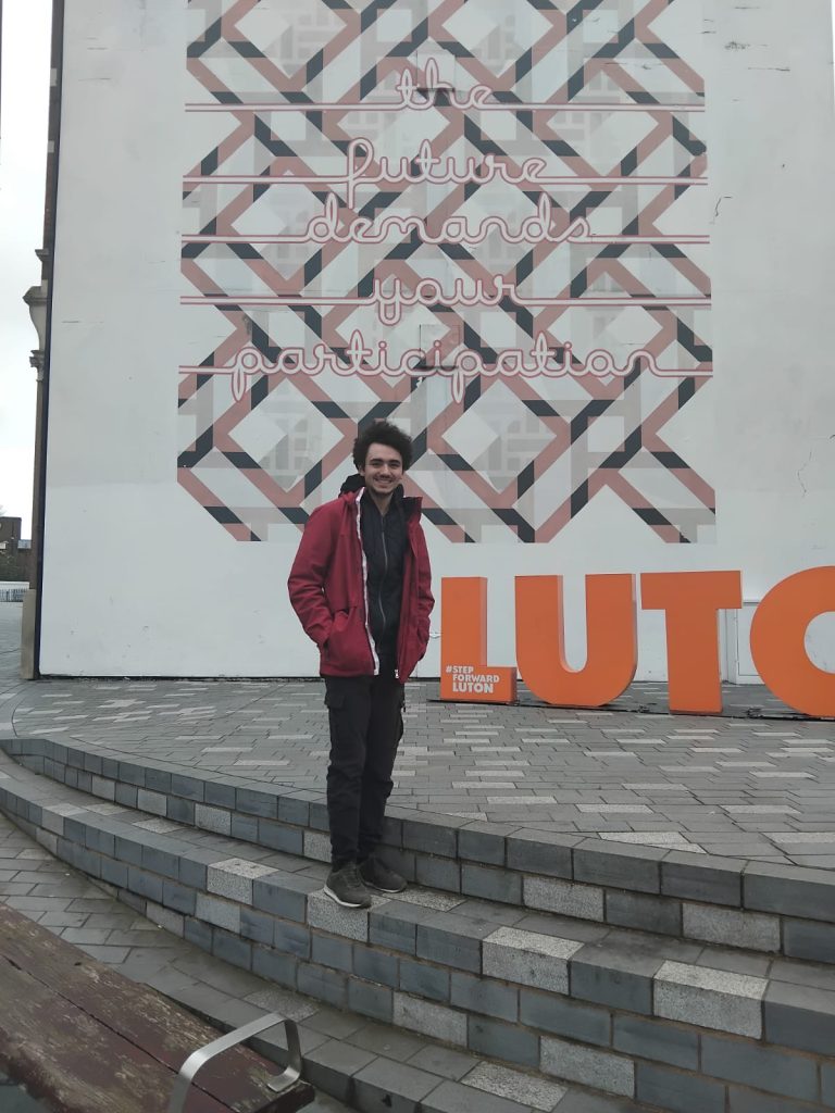 Manuel standing next to a mural in Luton with an orange letter sign saying LUTON.