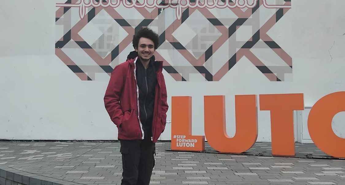 Manuel standing next to a mural in Luton with an orange letter sign saying LUTON.