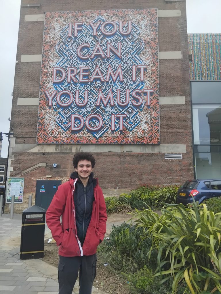 Manuel standing infront of a mural saying IF YOU CAN DREAM IT, YOU MUST DO IT