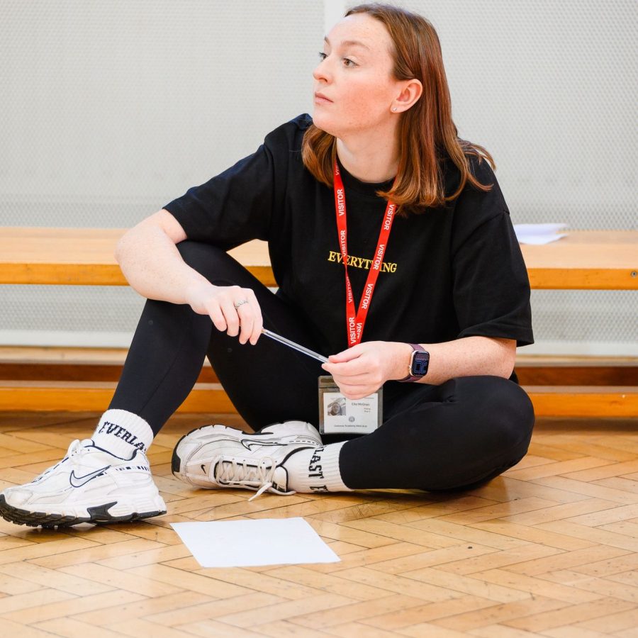 Ella sitting in a school hall setting with with legs crossed surrounded by notes on paper on a wooden floor.